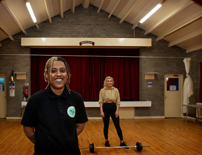 Image shows Yoko Jones in the foreground. Yoko is a black appearing woman wearing a black polo shirt. In the background is a cilent, white appearing, wearing sports wear, with a weighted bar in front.