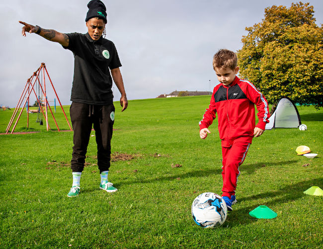 Image shows a young white appearing boy playing football, with Yoko Jones, a black appearing female, giving direction.