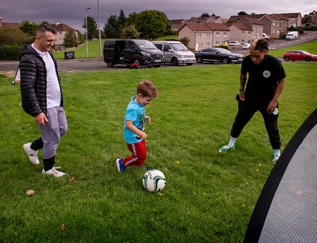 Image shows young boy and his father, both white appearing, playing football. Yoko jones, a black appearing woman giving instruction as the boy plays.