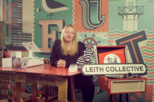 Portrait of Sara sitting at a table with till and car reader, next to sign reading 'Leith Collective'