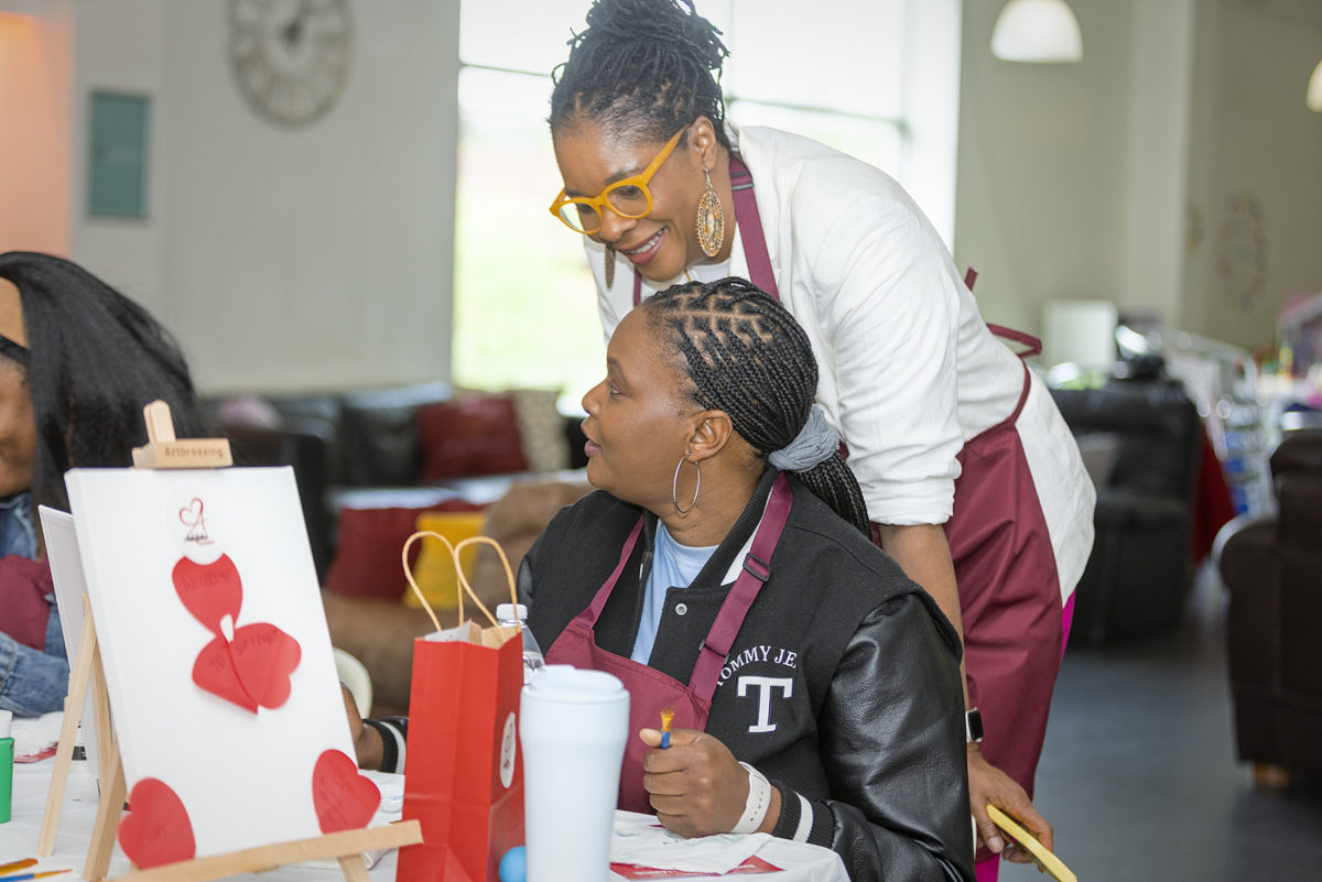 A woman stands with another person who is sitting, having a chat while the person sitting paints.