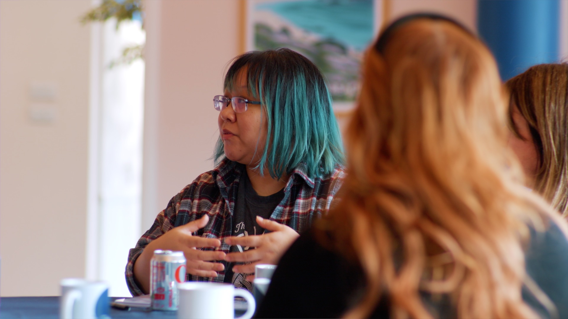 A person with blue hair and glasses speaks to others around a table during a meeting