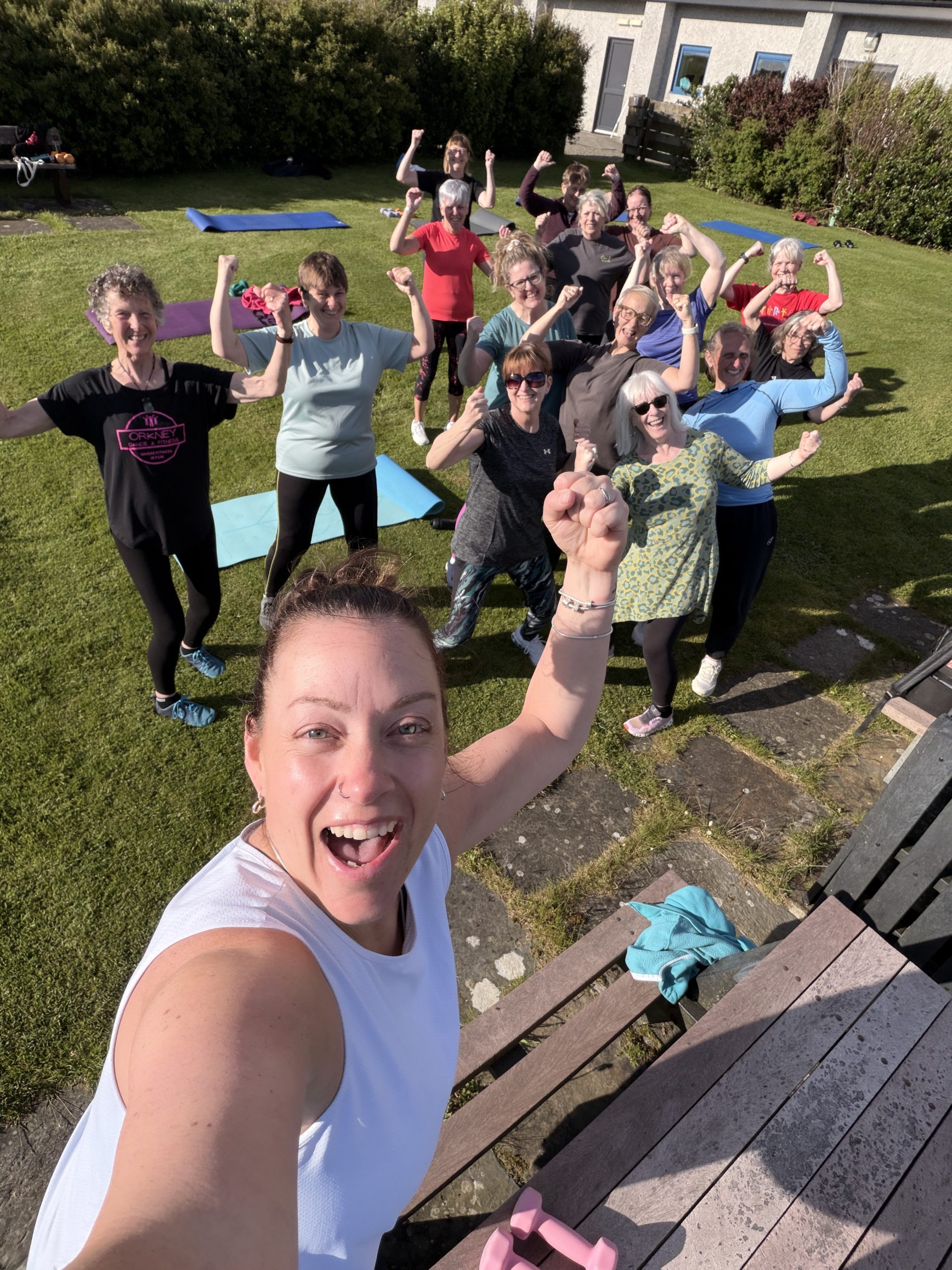 Donna alongside one of her Orkney Dance and Fitness classes. They are outside on grass and flexing their muscles to show their strength.